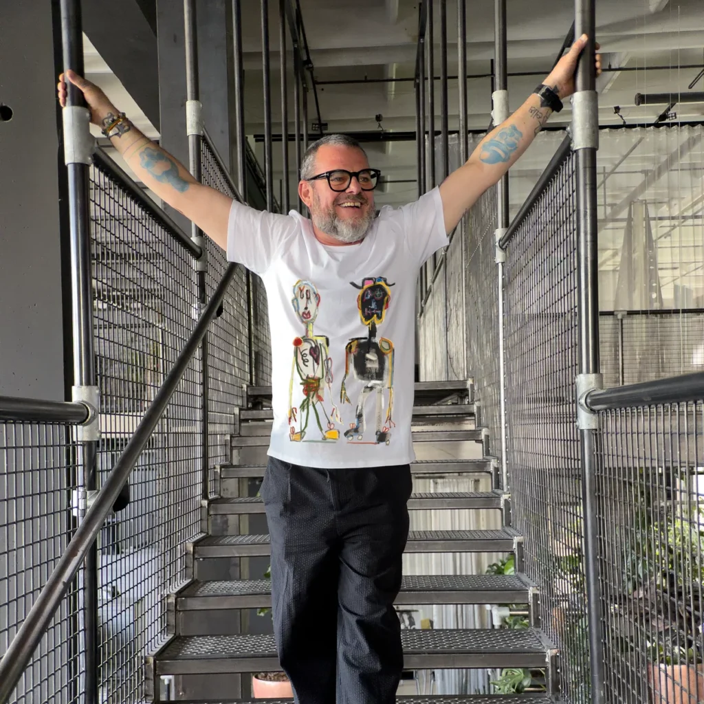 Accomplice with arms raised on an industrial staircase, wearing a printed t-shirt