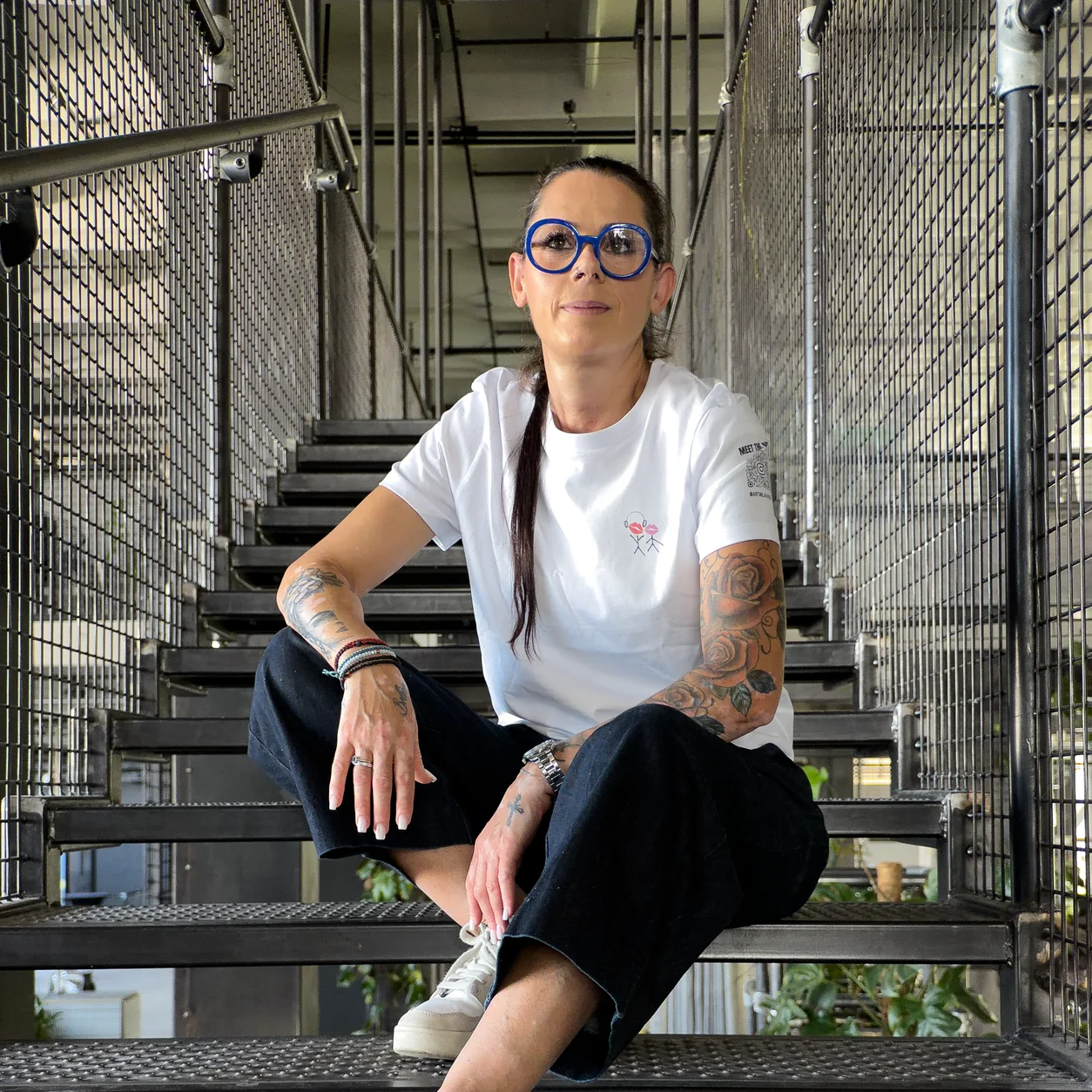 Accomplice sitting cross-legged on an industrial staircase in an artist-designed white t-shirt